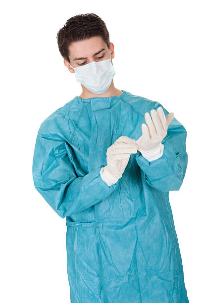 Surgeon putting on surgical gloves Young male surgeon in a mask and gown putting on disposable surgical gloves before going in to theatre isolated on white graduation clothing stock pictures, royalty-free photos & images