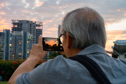 Elderly man uses smartphone to take pictures of city scenery at sunset