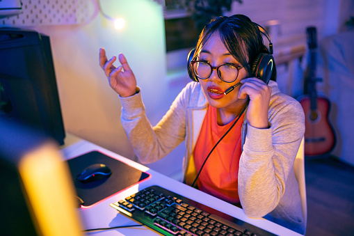 Modern young female gamer and streamer of Asian ethnicity, playing video games on the computer in her LED-lit room while having an chat with a follower during live stream