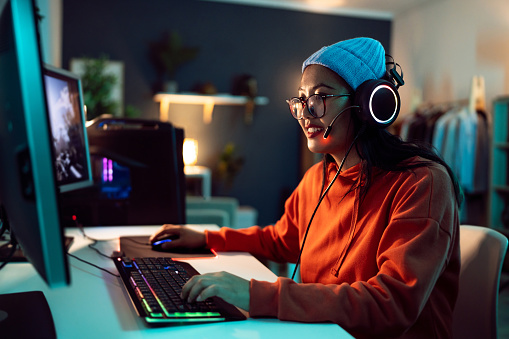 Young female gamer and streamer of Asian ethnicity, playing FPS video games on the computer in her LED-lit room