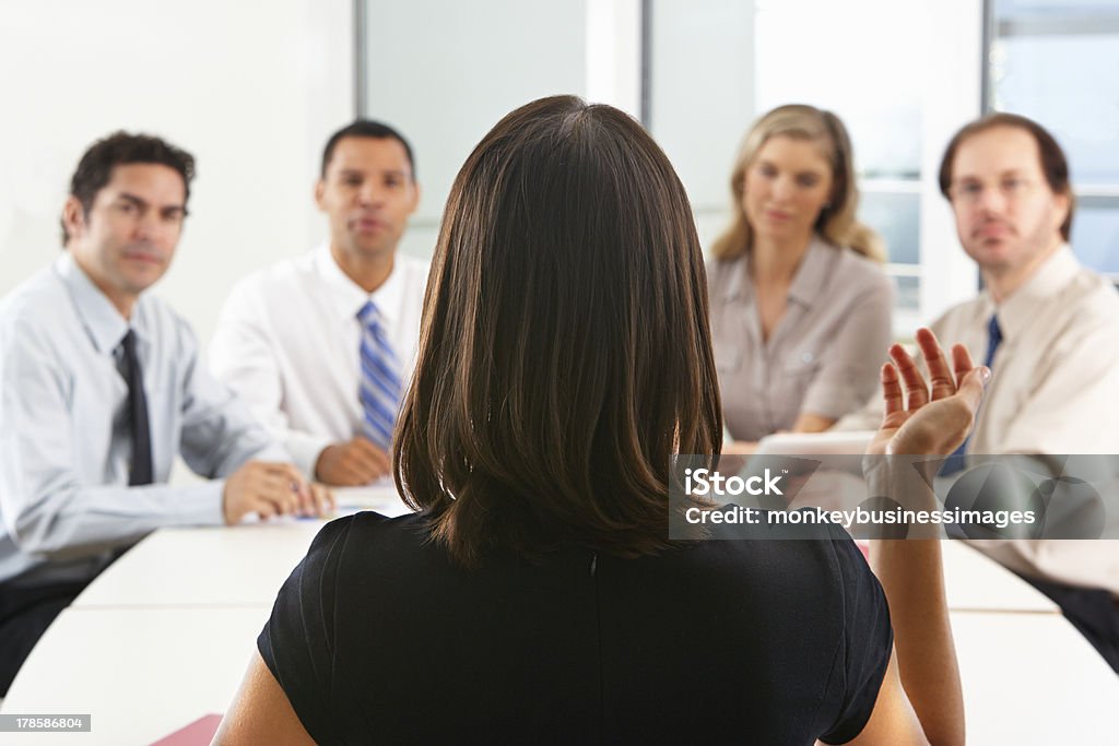 Group of people seated around boardroom table View From Behind As CEO Addresses Meeting In Boardroom Raising Arm Having A Discussion Meeting Stock Photo