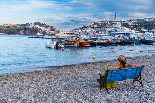 Woman with hat sits on a bench in the empty beach of Ponza Santa Maria overlooking the marina of the beautiful village of Ponza, Ponza Island, Pontine archipelago, Tyrrhenian sea, Latina province, Latium, Italy