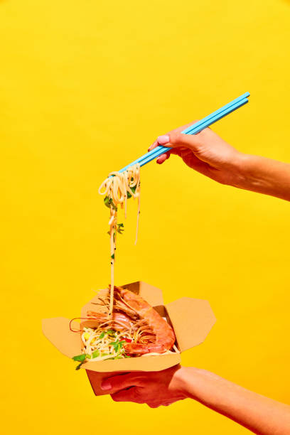Unrecognizable woman eating delicious hot WOK with shrimps with chopstick from paper box isolated yellow studio background