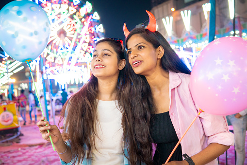 Two beautiful happy young female friends holding balloons and enjoying the local fair festival at evening.