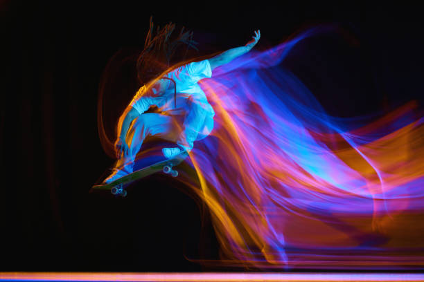 istock Young man, skateboarder with modern hairstyle practicing Ollie on skateboard with neon flashes of light isolated black background. 1785916950