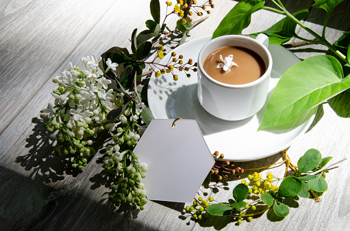Beautiful shadows with an coffee cup on a saucer with mockup an empty white card framed by branches of big green leaves, white lilac flowers and yellow small flowers