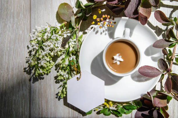 Beautiful shadows with an coffee cup on a saucer with mockup an empty white card framed by branches of burgundy and green leaves, white lilac flowers and yellow small flowers