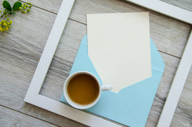Flatlay gray natural wooden background with square white wooden frame and blue envelope white postcard copyspace, small yellow flowers and green leaves on a branch