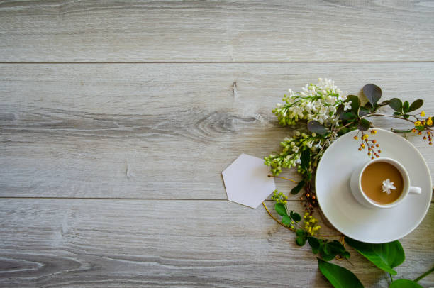 istock Flatlay with an coffee cup on a saucer with mockup an empty white card framed by branches of green leaves, white lilac flowers and yellow small flowers 1785920838