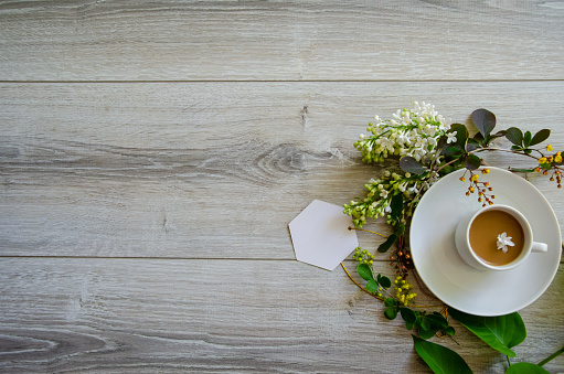 Flatlay with an coffee cup on a saucer with mockup an empty white card framed by branches of green leaves, white lilac flowers and yellow small flowers