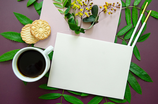 Flatlay close-up pink mockup envelope filled with yellow small flowers and small leaves with a spring green leaves around, White card mockup and a cup of coffee, cookie, dark purple background