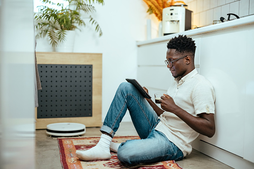 ​Man sits on the kitchen floor while a robot vacuum cleaner vacuums for him