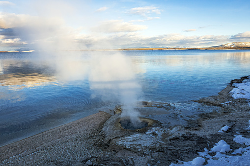 Yellowstone Lake at sunset with a thermal geyser at the edge of the shore with calm water reflecting the surrounding winter landscape in winter.