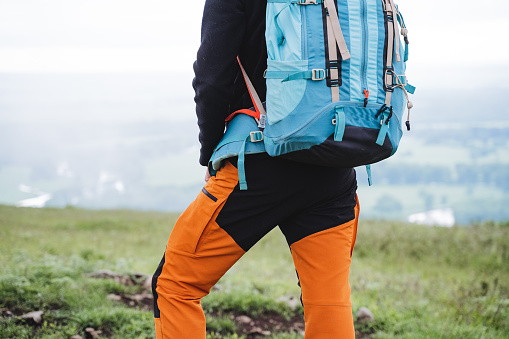 An element of sports equipment is a tourist backpack for hiking in the mountains, a person stands on top of a mountain, a view from behind a tourist in pants, equipment for a hike. High quality photo