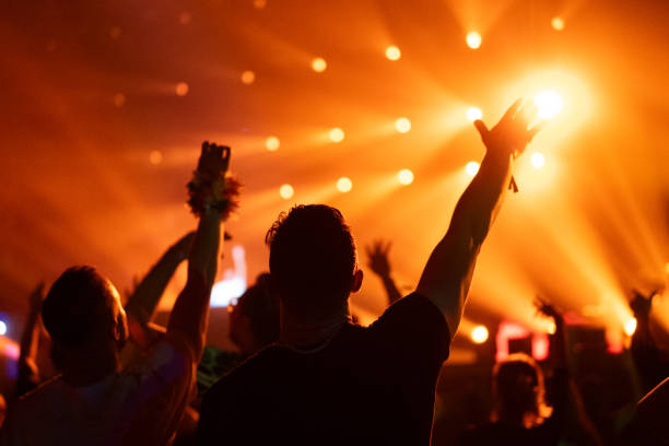 istock Crowd of people attending a musical performance. Audience dancing illuminated by orange light beams coming from stage 1786025255