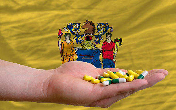 holding pills in hand front of new jersey flag man holding capsules in front of complete wavy american state flag of new jersey symbolizing health, medicine, cure, vitamines and healthy life jerseyan stock pictures, royalty-free photos & images