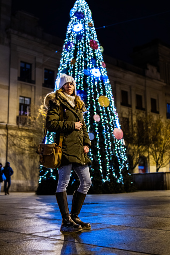 Young girl with hat, coat, bag and high boots, Caucasian origin stands in front of a Christmas tree.