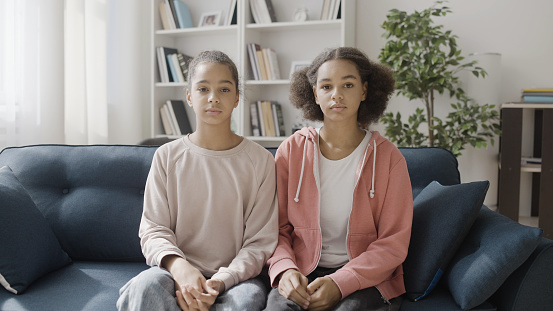 Two African American sisters sitting on sofa and seriously looking at camera