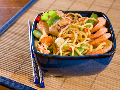 Noodles with fish,vegetables, chili peppers and coriander in a blue bowl on a placemat with chopsticks.