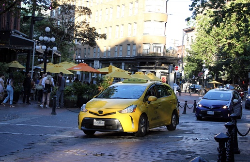 Vancouver, Canada – July 28, 2023: A yellow taxi car is seen driving down a busy city street, with people walking on the sidewalk in the foreground