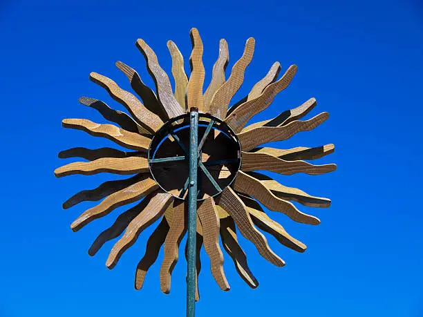 A wood whirlygig in the form of a sun with radiating rays outlined against a bright blue sky.