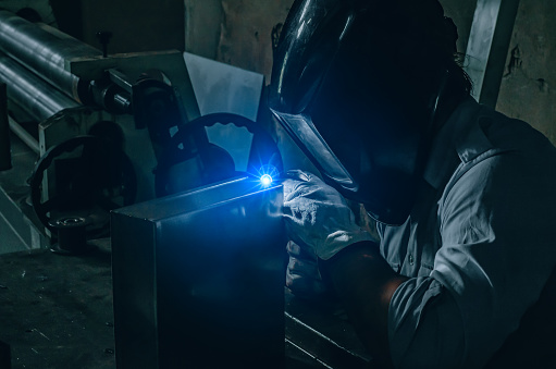 A welder in a protective mask welds a metal box in a locksmith's workshop. Bright light of argon welding