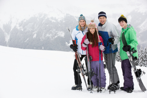 Family On Ski Holiday In Mountains Holding Skis And Poles Smiling To Camera