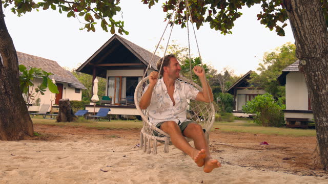 Slow Motion: Man swinging on an hanging chair in tropical beach settings, beach holiday vacation concept.