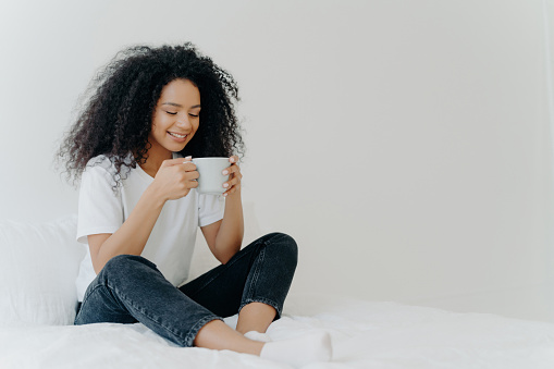 Smiling young woman enjoying a warm drink, sitting cozily on a bed with a relaxed pose