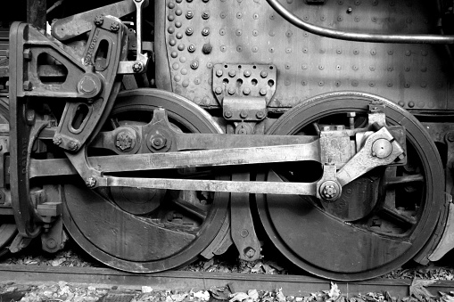 This grainy black and white image shows the train wheels and power linkages of an old locomotive engine parked on railroad tracks.  Large rivets holding the steel plating on the engine are seen.  This vintage image is a reflection of America's industrial revolution and evokes a by-gone era.  The season is fall and dry leaves can be seen along the bottom of the photo.   The photo was taken in the early fall in the town of  Snoqualmie, Washington.