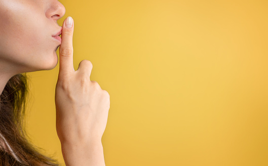 Calm young caucasian lady puts finger to lips, making shh sign, isolated on yellow studio background, profile, close up. Secret gesture, gossip, good news, ad and offer, human emotions