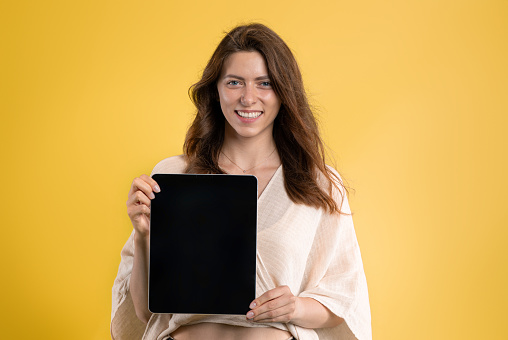 Happy young caucasian woman in casual show tablet with blank screen, isolated on yellow studio background. Sale with device presentation, recommendation app, ad and offer, good news