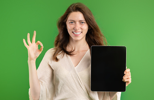 Cheerful young european lady show ok sign with hand and tablet with empty screen isolated on green studio background. Blog, website recommendation sale and app, ad and offer