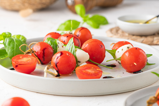 Caprese skewers with tomatoes, mozzarella balls, basil and spices on white table, closeup.  Italian homemade food and a healthy diet concept.