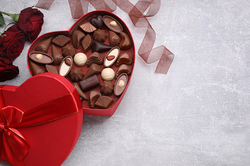 Heart shaped box with delicious chocolate candies, roses and ribbon on light grey table, flat lay. Space for text