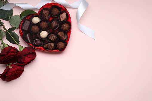 Heart shaped box with delicious chocolate candies, roses and ribbon on pink background, flat lay. Space for text