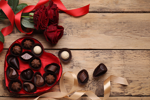 Heart shaped box with delicious chocolate candies, roses and ribbon on wooden table, flat lay. Space for text