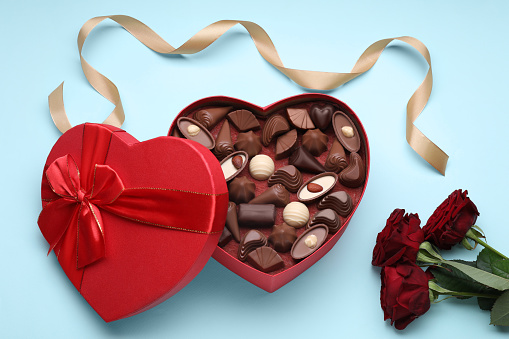 Heart shaped box with delicious chocolate candies, roses and ribbon on light blue background, flat lay