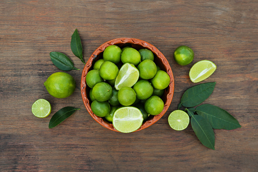 Whole and cut fresh ripe limes in bowl on wooden table, flat lay