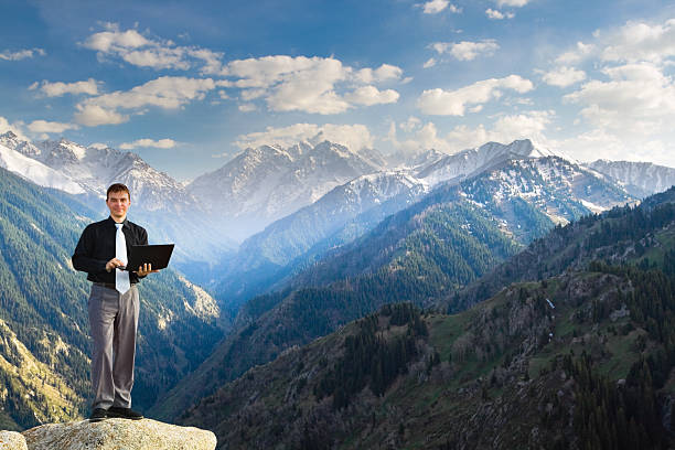 Young businessman using his laptop on the mountain top "Image of a businessman standing on the top of the mountain, using a laptop and looking at the camera happy with his success. In the background a beautiful panorama of the valley with high peaks." environment computer cloud leadership stock pictures, royalty-free photos & images