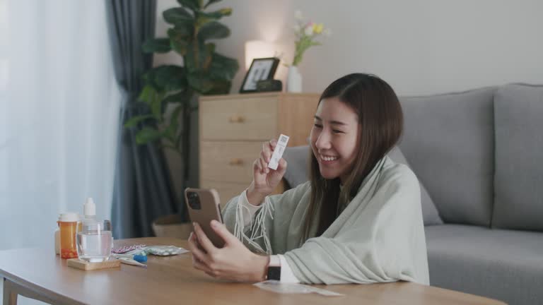 Smiling an Asian woman showing rapid antigen test kit result negative to friends and family via video call on phone during the pandemic of disease, COVID-19, and coronavirus at home.