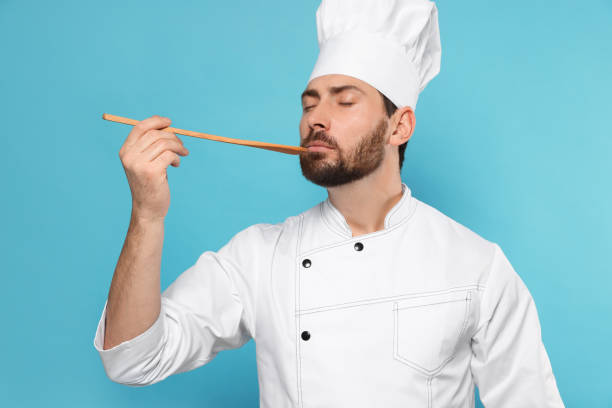 istock Mature male chef with spoon tasting dish on light blue background 1786364055