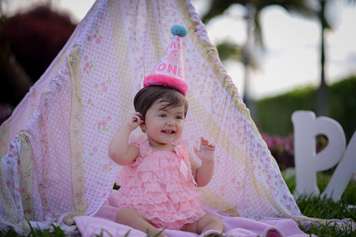 Portrait of one-year-old girl sitting in pink decorative tent tent and party cap as she smiles