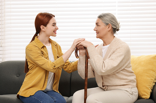 Caregiver and senior woman with walking cane sitting on sofa at home