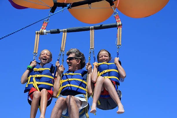 Happy Family Parasailing Father and Twin Daughters Parasailing Against a Blue Summer Sky. Shallow dof with focus on father's face. parasailing stock pictures, royalty-free photos & images