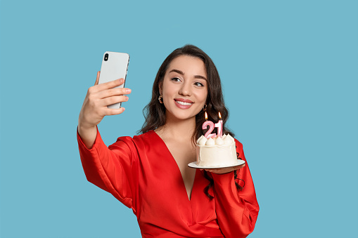 Coming of age party - 21st birthday. Woman holding cake with number shaped candles and taking selfie against light blue background