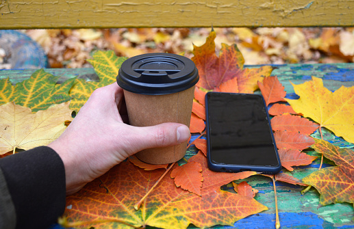 a man's hand holds a glass of coffee against a background of autumn leaves, a smartphone lies nearby. outdoor walks and an active lifestyle. communication by phone and via the Internet