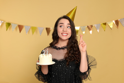 Coming of age party - 21st birthday. Smiling woman holding delicious cake with number shaped candles and crossing fingers on beige background