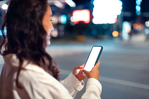 Young woman using smartphone with blank screen, standing at night city street near road, using new mobile app for navigating in megalopolis, mockup, free space