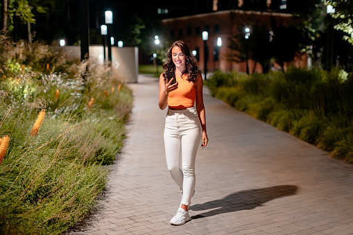 Full length shot of young european woman in casual outfit walking on the city street at night and using cellphone, texting or browsing online maps app, free space